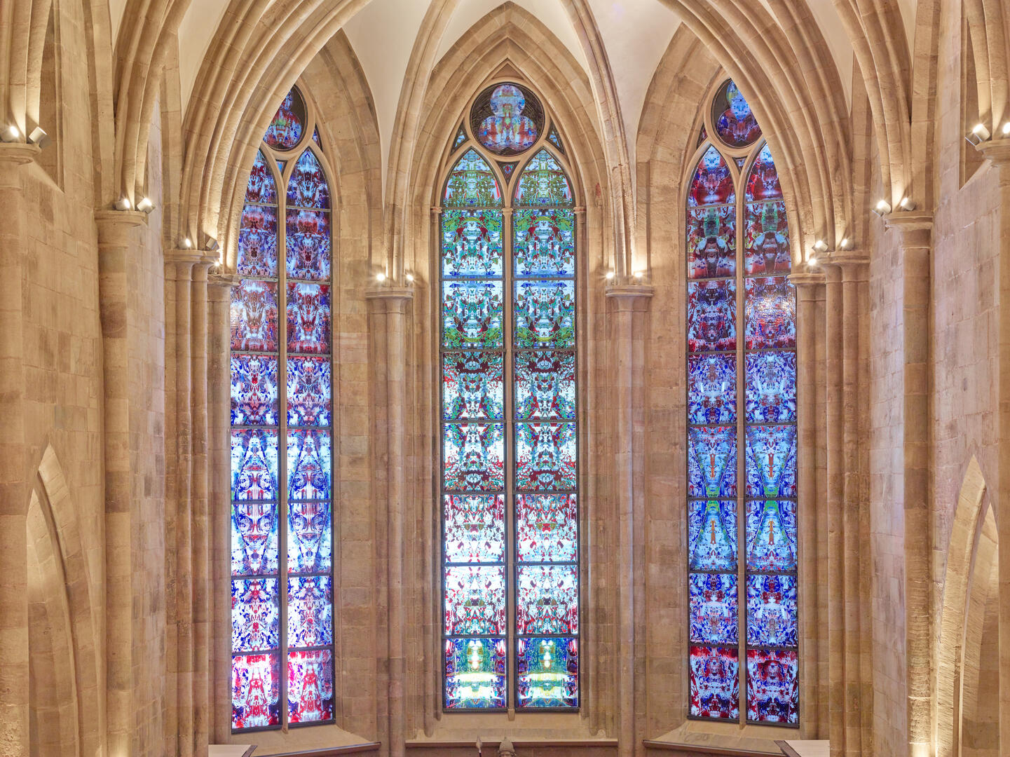 The three Richter choir windows at Tholey Abbey, with designs like a Persian rug or kaleidescope; the far left window mostly pinks, blues and purples; the middle of greens, reds and blues; the right of similar coloring to the left, but deeper hues, and bits of green