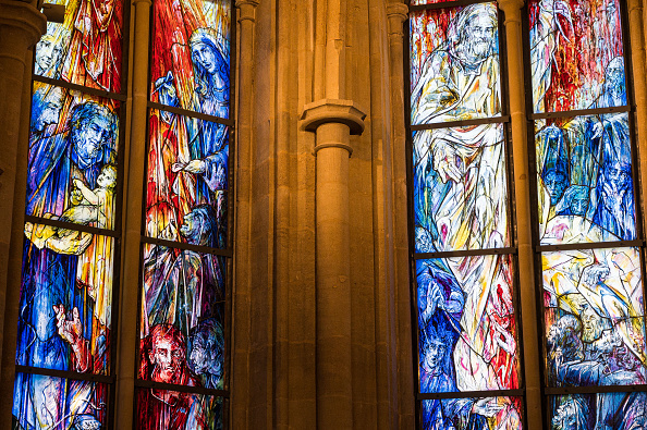 Two windows in Tholey Abbey; On the left, a Christmas image of Joseph holding baby Jesus near Mary in reds, blues and yellows; on the right, a Pentecost image of Jesus in white and yellow, the disciples in red and blue