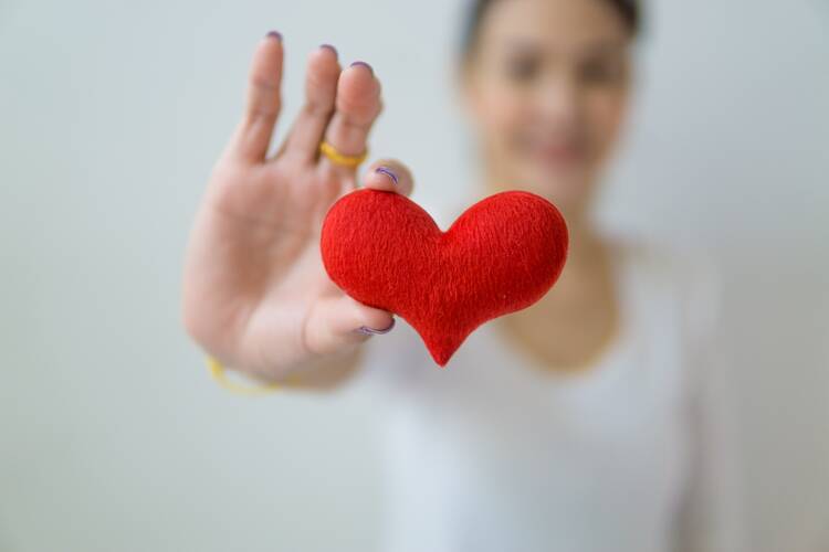 A woman, blurred out in the background, holds a red heart made of felt-like material in the foreground of the photo
