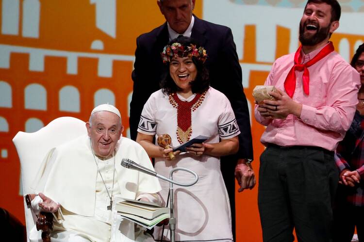 Pope Francis, seated, smiles as he speaks into a microphone, flanked by a laughing man and woman
