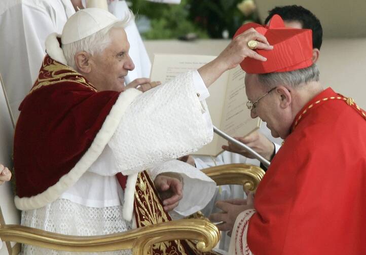 A cardinal dressed in red kneels in front of Pope Benedict XVI, seated and dressed in white