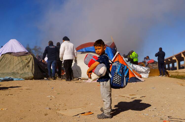 a child stands holding some items of clothing as police move people from a camp in the background behind him