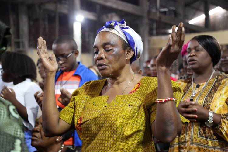a congolese woman in a yellow dress and blue and white headscarf prays with hands uplifted and eyes closed