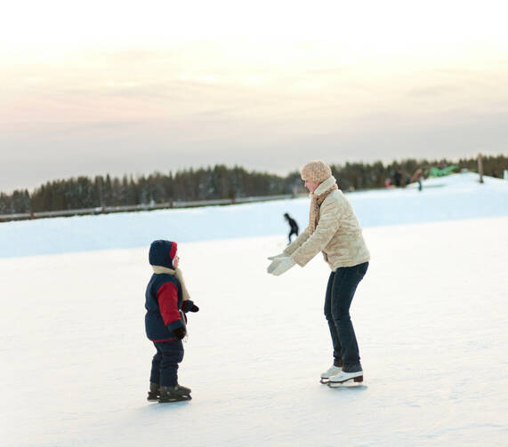 woman wearing a white coat and hat coaches a student wearing a red coat while both are ice skating. the woman reaches out to the child