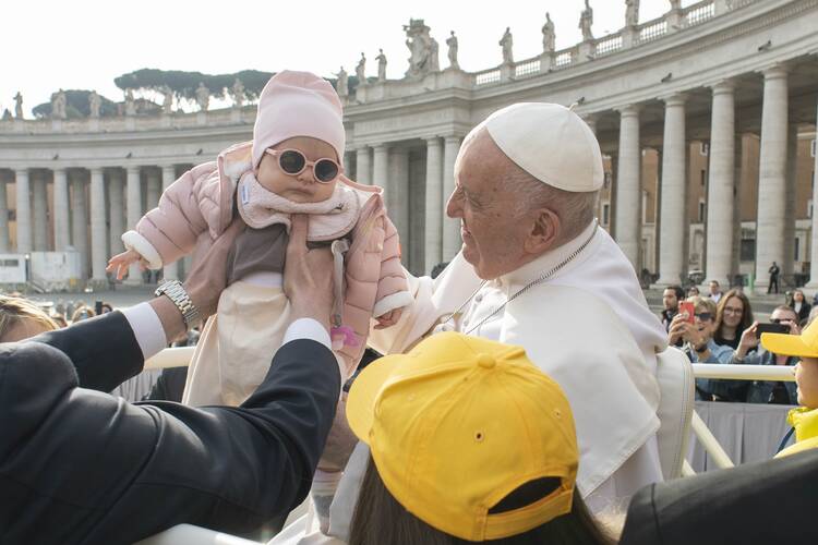 pope francis at left greets a baby wearing a light pink hat and jacket and sunglasses