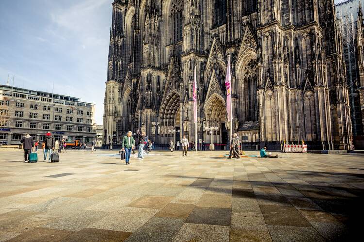 Wide shot on a cloudy day of the cathedral in Cologne, Germany, with people walking outside on the sidewalk.