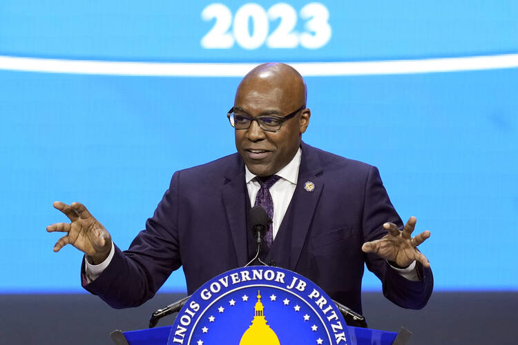 illinois attorney general kwame raoul, a bald black man wearing a suit, gestures while speaking in front of a bright blue wall with the seal of illinois in front of him