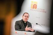 bishop georg bätzing sits in front of a microphone at the conference of the german bishops, he wears his priest clothing and is gesturing with his hands