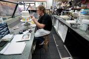 jack phillips sits in his cake shop decorating a white cake, he is wearing a black shirt and has cakes on the table behind him