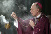 A man in red robes and a bishop's hat swings incense in a gold burner