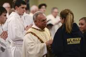 A priest in white robes speaks with a young girl in a jacket that reads "Newtown Lacrosse"