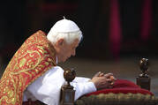 pope benedict xvi kneels on a kneeler with a red cover, he is wearing white and a red mantel