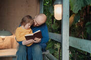A man reads to a toddler sitting on his lap on a porch