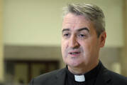 a priest wears a collar and talks in front of a beige background, he has gray hair and dark eyebrows