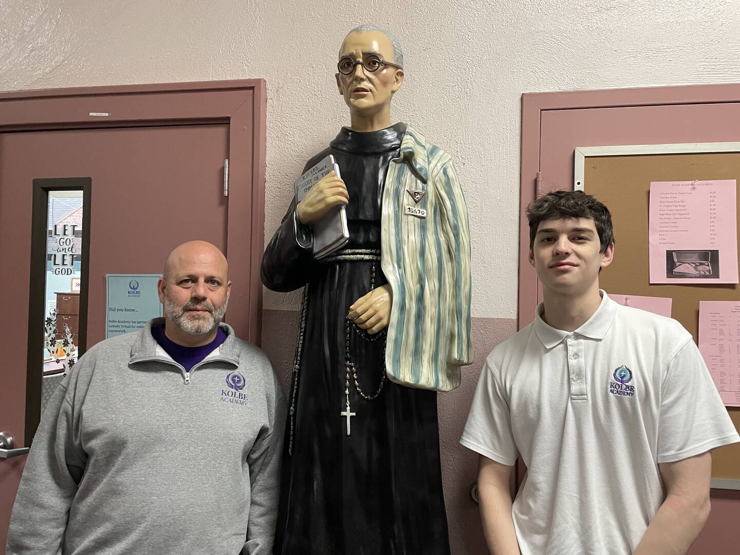 an older man and a teenager stand in front of a statue of maximilian kolbe at the school named for him