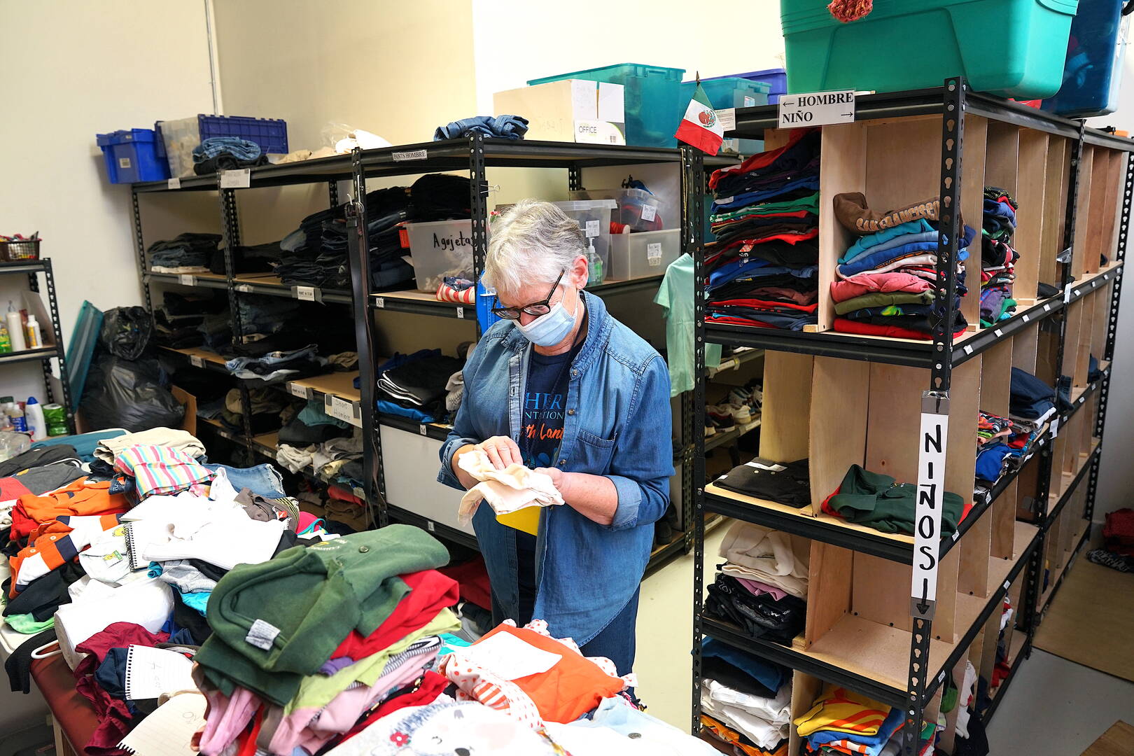 a woman stands sorting clothing for migrants, racks behind her are for men and boys