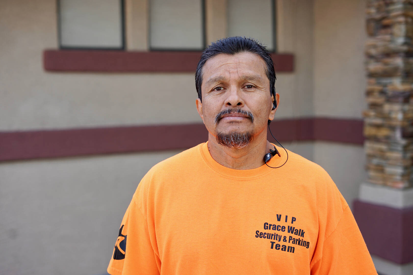 a hispanic man stands in front of a building, he wears a mustache and an orange shirt