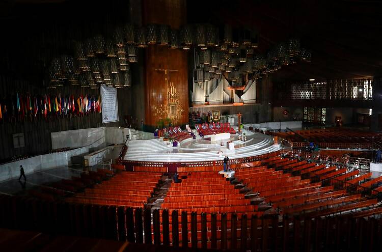 A large sanctuary with reddish brown pews is empty, from the distance, Mexican Cardinal Carlos Aguiar Retes can be seen standing on a white square platform at the end of the pews, preaching to the empty space. A large organ and array of flag are on the wall behind the altar.
