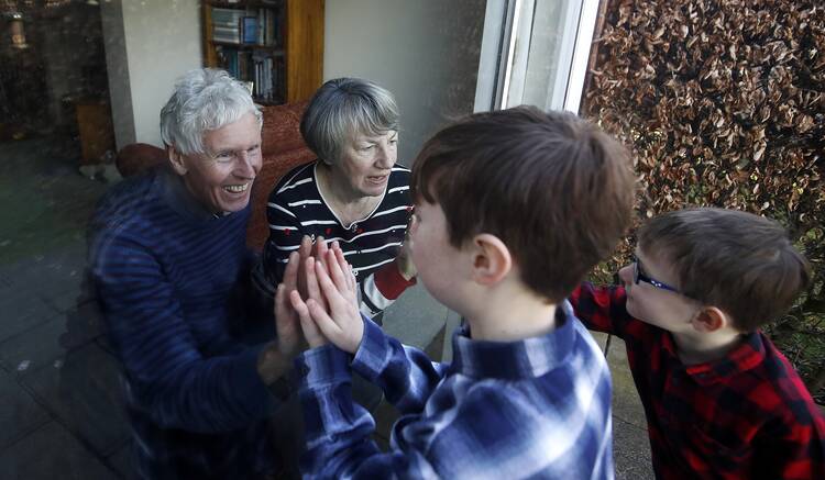 Two grandparents, an older white woman and white man with grey hair, sit behind a glass or plastic screen, touching hands with two of their grandchildren, a boy and a girl, who from their profile are also light-skinned.