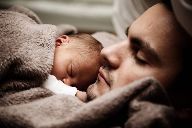 A newborn baby rests on a father's chest nuzzled up to his chin, both have eyes closed, are white; the father has dark brows and a bit of a mustache growing in; they are wrapped in a light brown blanket