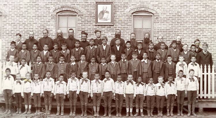 Jesuits and boys under picture of St. Ignatius at the Holy Rosary Mission circa 1880-1900. Courtesy of Marquette University, Raynor Memorial Libraries and Holy Rosary Mission – Red Cloud Indian School Records, ID: MUA_HRM_RCIS_02937.