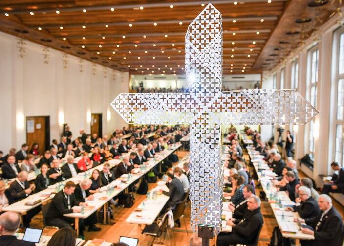 A metallic, contemporary-styled cross in the foreground while German bishops sit at long tables in the background.