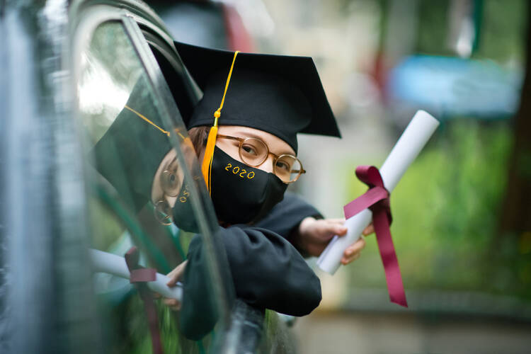 A college graduate wearing a mask leans out of a car window holding her diploma.