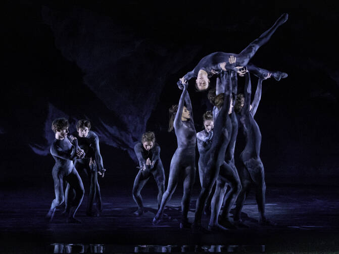 Five artists of The Royal Ballet in Wayne McGregor’s “The Dante Project” hold up another dancer above them under blue stage lights.