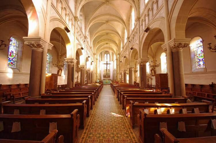 Empty pews in an ornate, sunlit Catholic Church with a cream-colored ceiling.