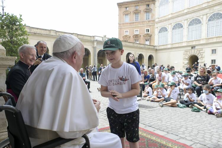 The Pope, seated before a crowd, greets a young boy in a green hat.