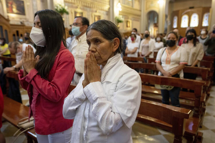 Two women stand with hands clasped in prayer in the foremost pew of a church