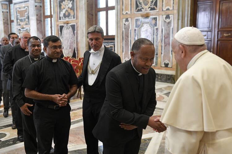 The pope shakes hands with a priest. Behind the priest, others wait in line.