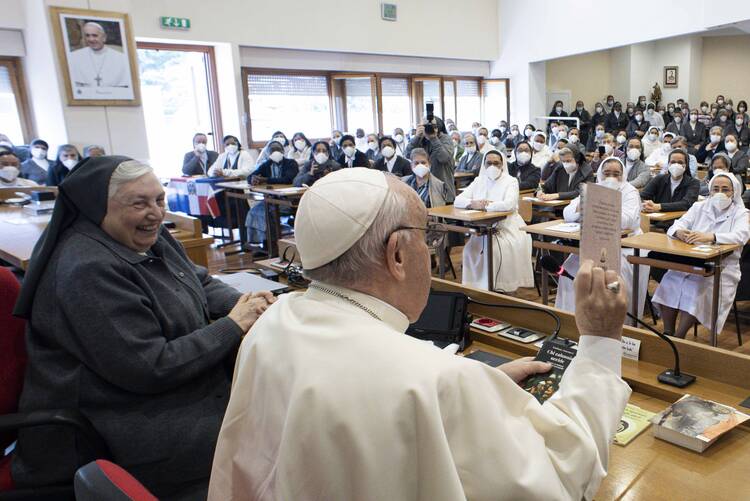 A smiling religious sister in habit sits to the left of Pope Francis before a crowd of sisters