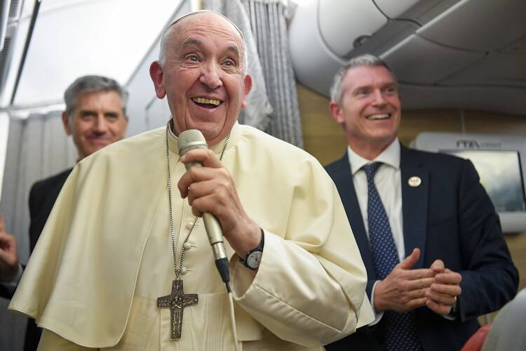 The pope, holding a microphone and smiling, receives questions from reporters on an airplane.