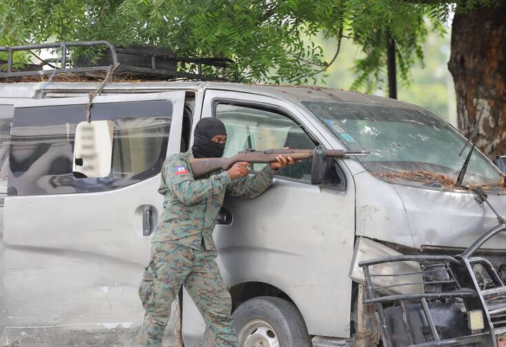 A soldier wearing a ski mask and holding a gun hides behind a broken-down car.