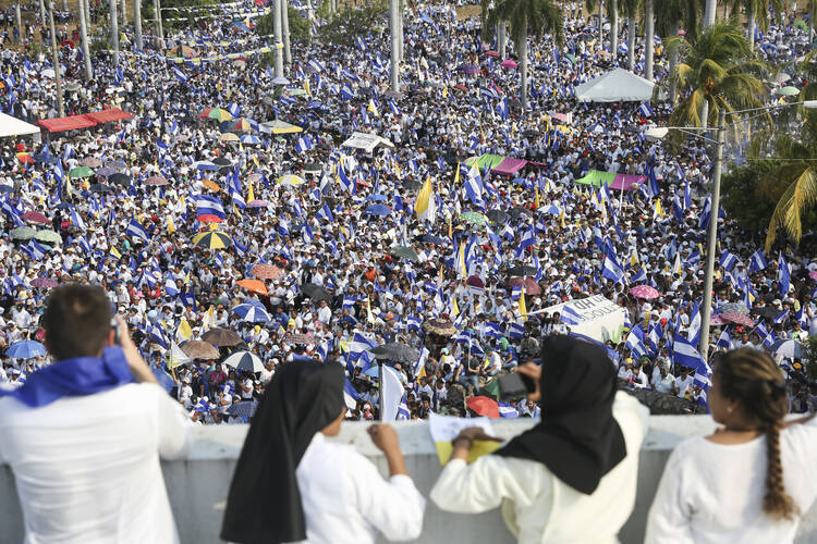 Two women in habit look over a ledge at a large crowd of people holding flags