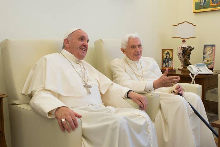 pope francis sits on a chair next to retired pope benedict. both are wearing white and smiling