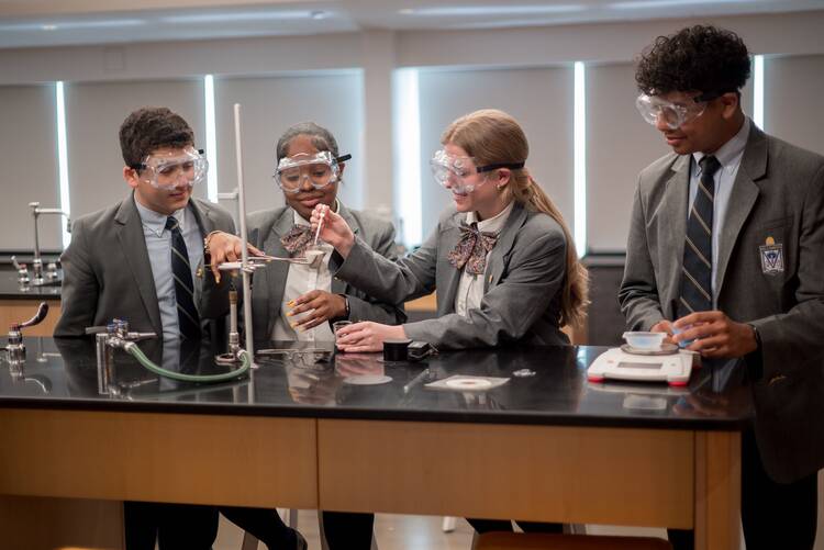 students at Kellenberg High do STEM science project in a classroom with lab equipment, all four students are wearing gray blazers and safety goggles
