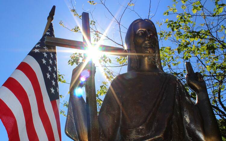 an american flag stands to the left next to a crucifix with light coming from behind it, a bronze statue of Jesus is to the right