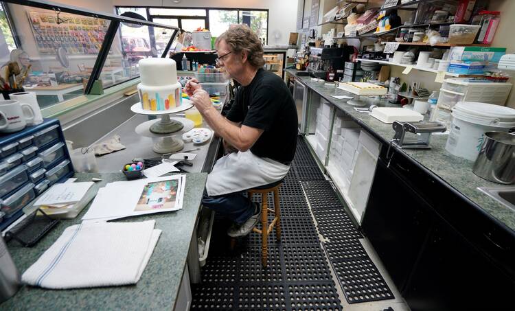 jack phillips sits in his cake shop decorating a white cake, he is wearing a black shirt and has cakes on the table behind him