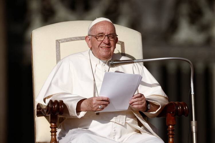 pope francis sits in his chair and smiles, he wears glasses and is holding a paper