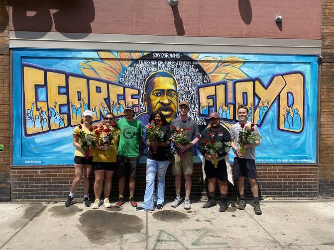 seven college students in regular clothes stand in front of a graffiti mural of george floyd with his face and name, in minneapolis