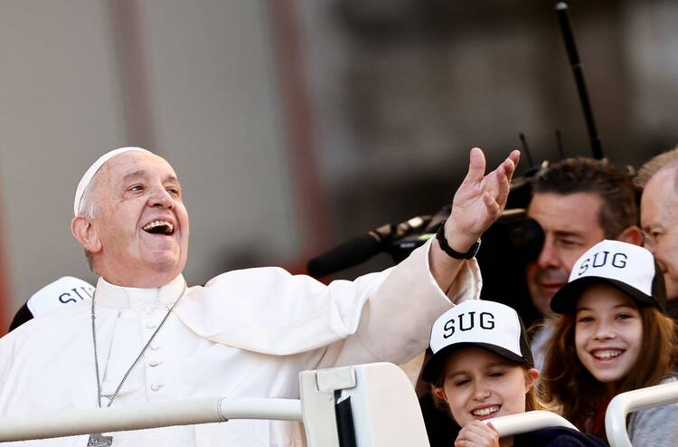 pope francis greets people with his arm out and a big smile on his face, he sits in his popemobile and the crowds are behind and below him