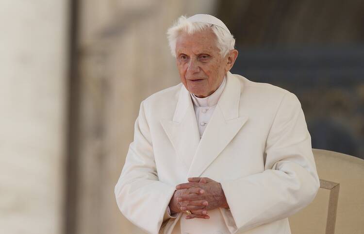 pope benedict xvi stands with hands clasped wearing white with a blurred background behind him