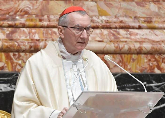 A man in white robes wearing the red cap of a cardinal speaks into a microphone