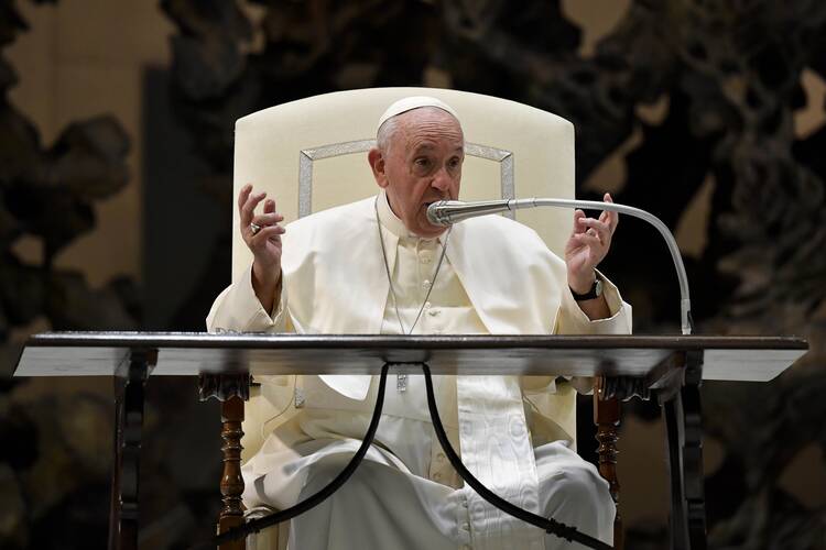 pope francis sits inside on a white chair with a microphone in front of him and his hands in front of him gesturing while speaking. he is wearing his usual white clothing
