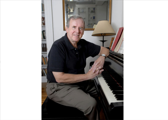 thomas cahill sits in his new york apartment in a 2006 photo, he is wearing a black shirt and brown pants and rests his arm on a piano. there is a lamp behind him