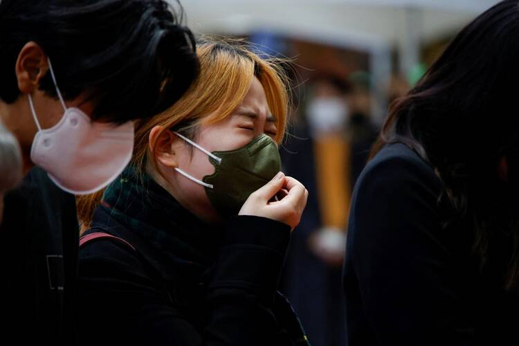 a woman cries at a group memorial for the victims of the stampede. she has dyed orange-red hair and is wearing a green mask and is covering her mouth in sorrow