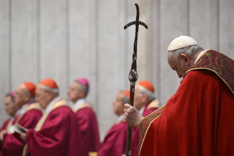pope francis to the right, wearing red vestments, bows his head while praying. he holds a metal crucifix staff in one hand. in the background are some cardinals sitting also in red vestments