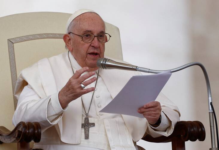 pope francis sits at his chair holding a paper and gesturing with his right hand, he is wearing glasses